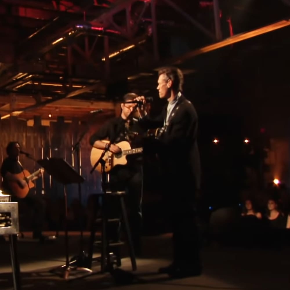 Musical performance in an indoor venue with multiple adult male performers, one holding a guitar and singing into a microphone.