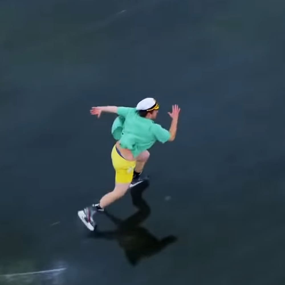 Young adult male in a dance studio wearing a green button-down shirt, yellow tight-fitting shorts, white cap with yellow band, and white sneakers with red accents; dark flooring suggests an indoor dance studio environment.