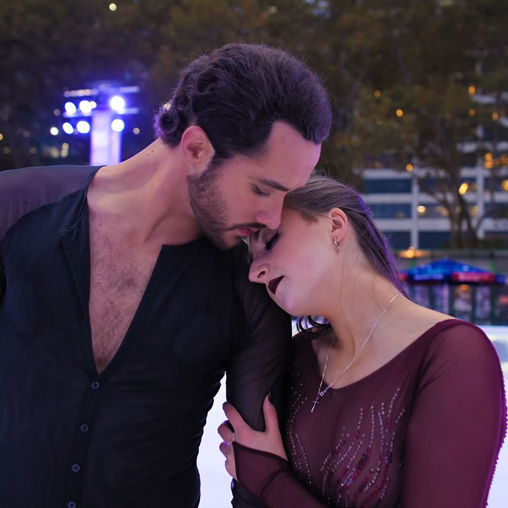 A young adult male and female couple outdoors at night. The man wears a black button-up shirt, while the woman dons a maroon, sheer dress adorned with small decorative beads or sequins, silver jewelry, against an illuminated blue background suggesting an ice rink setting.