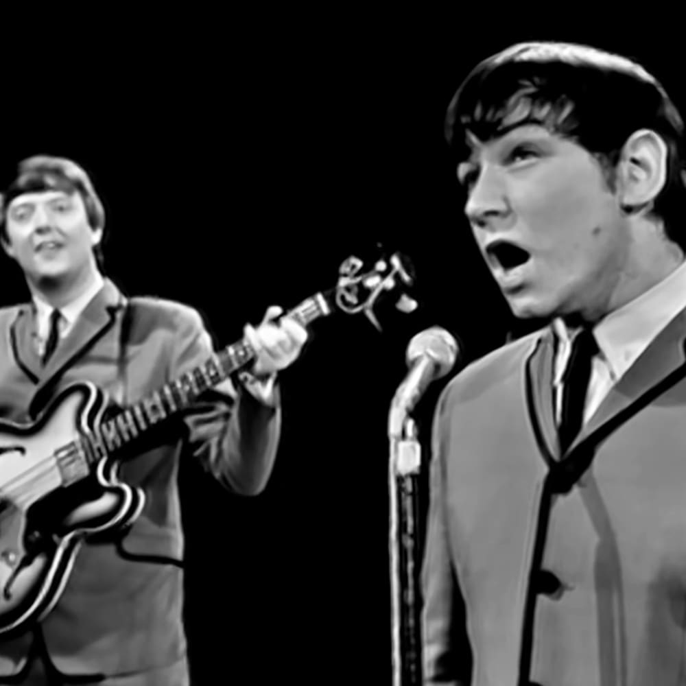 A musical performance scene with two young adult male performers wearing light-colored suits with distinctive dark tie designs against a dark background.