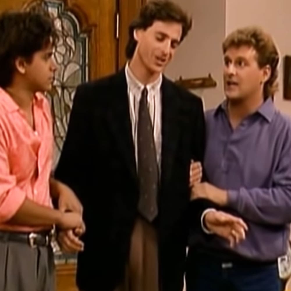 A young adult male wearing a dark suit jacket and patterned tie stands center, flanked by a child/teenage boy on the left and an adult male on the right in an indoor setting with wooden decor.