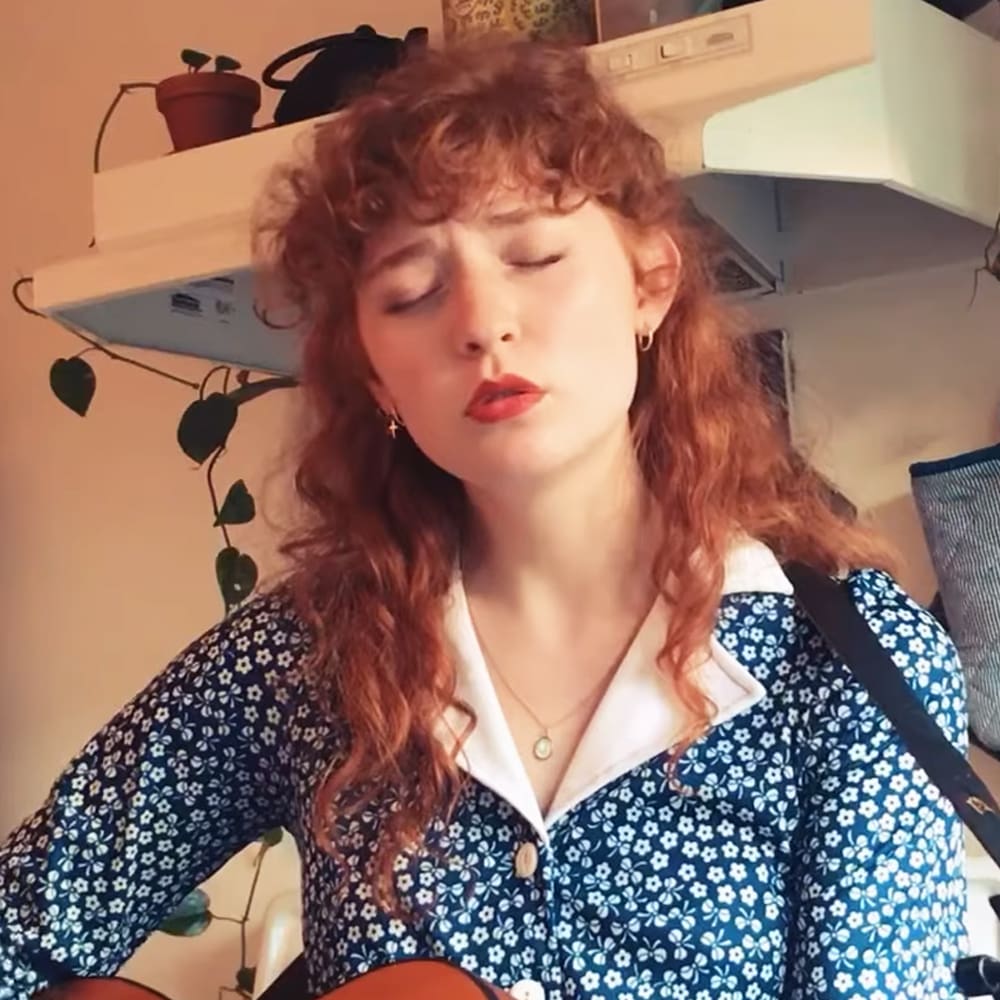 A young adult female in a relaxed, casual fit floral-patterned blue and white shirt with long sleeves and a white collar, wearing gold jewelry including an oval pendant necklace and small hoop earrings. She is indoors in what appears to be a home setting, surrounded by shelves of plants and other items, with a guitar strap visible over her shoulder.