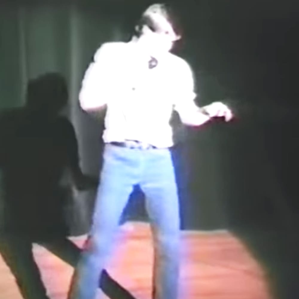 A young adult male wearing a white long-sleeve shirt with buttons and blue jeans stands indoors under dim lighting.