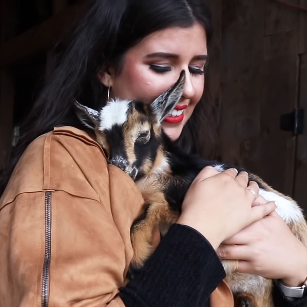 A young adult female holding a German Shepherd in an indoor barn setting with wooden walls and dim lighting, wearing a brown leather jacket and red lipstick.