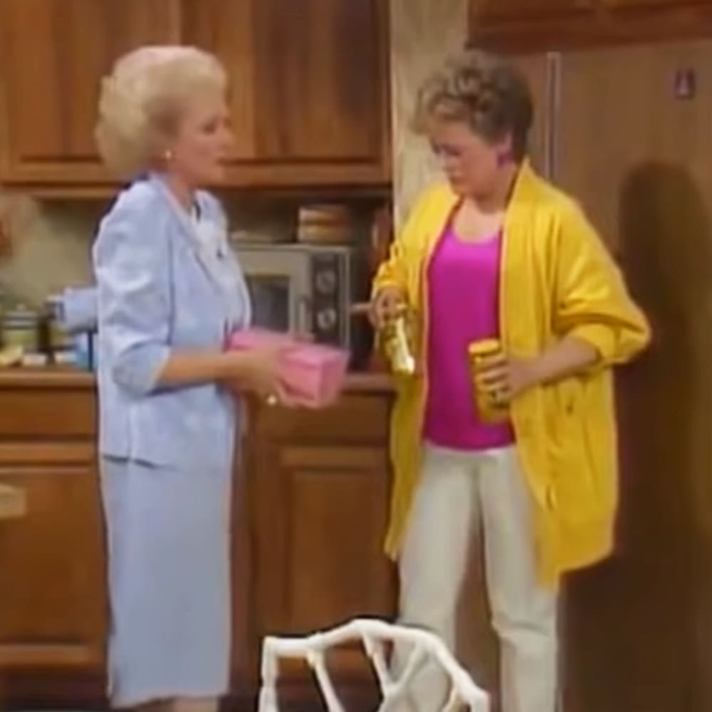 Indoor kitchen scene with two adults, one in a light blue dress and the other in a yellow cardigan. A white dog stands near them, surrounded by wooden cabinetry and appliances.