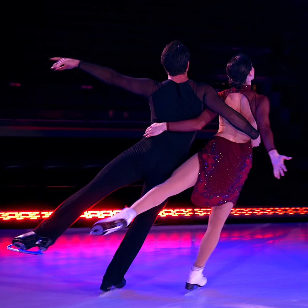 A figure skating pair on an indoor ice rink with a young adult male skater wearing black pants, a fitted black top, white boots, and a young adult female skater in a red sequined dress, empire waist, above-knee skirt, thin straps, and white boots. The performance is set against blurred stage lights suggesting artificial lighting for an indoor arena.