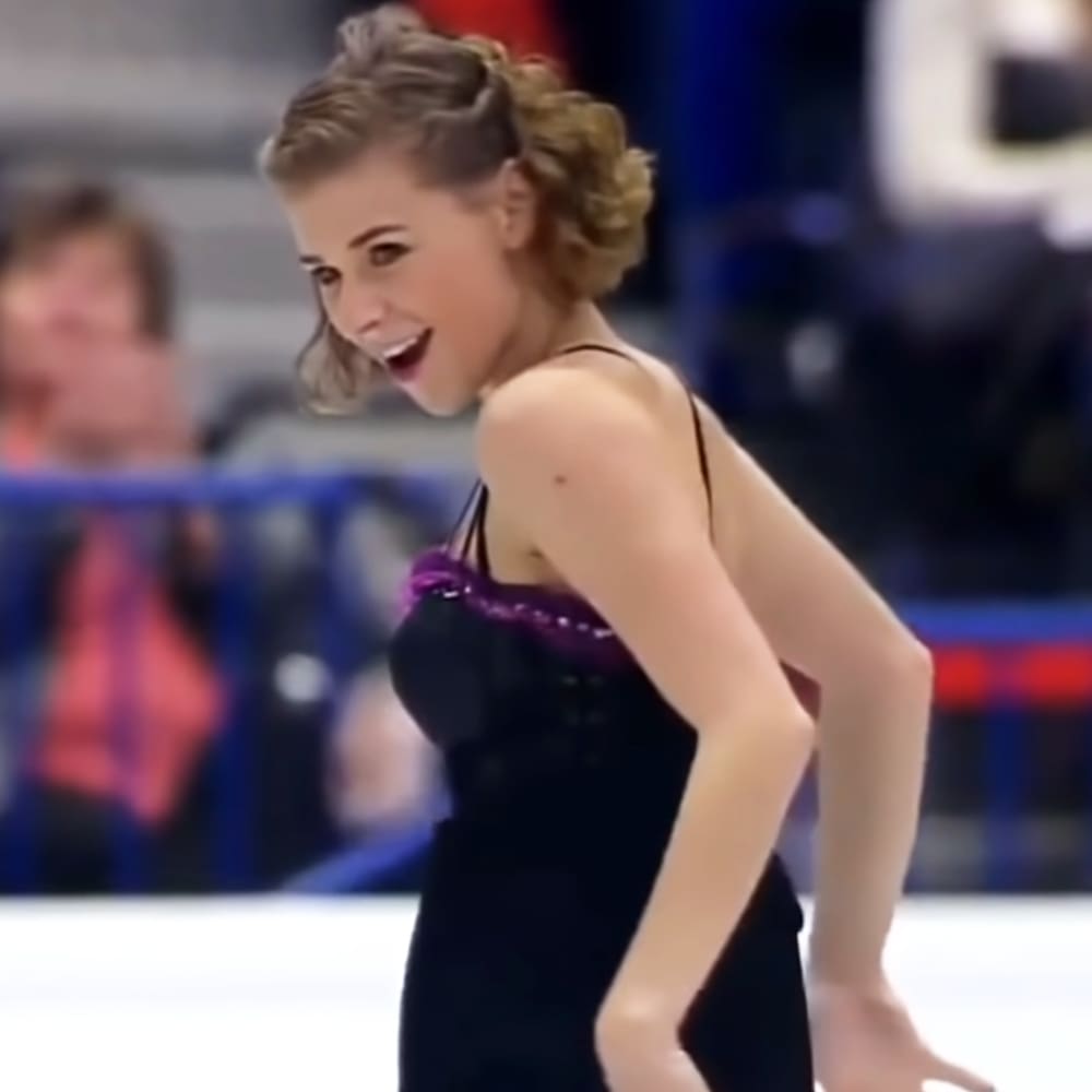 A young adult female figure skater in a black leotard with purple ribbon, skating on an indoor ice rink under bright even lighting.