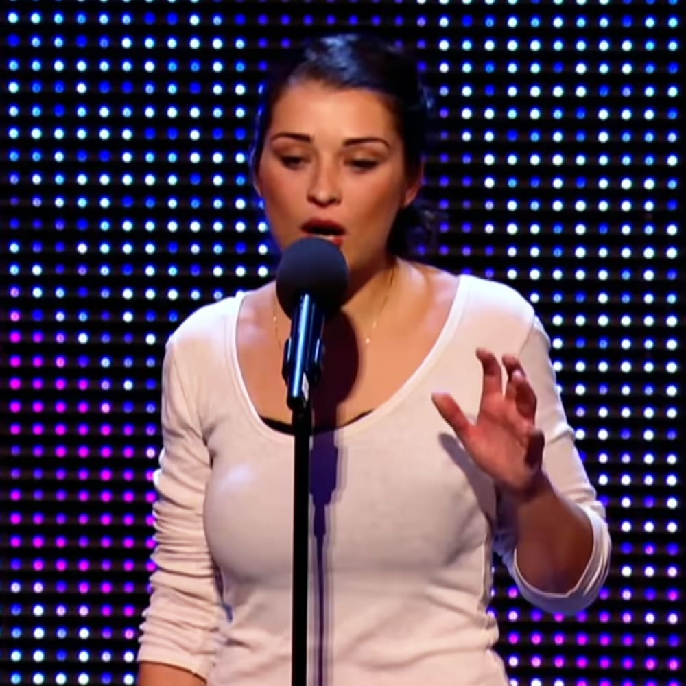 A young adult female in a white long-sleeved top performs at an indoor event with stage lighting and microphone setup.