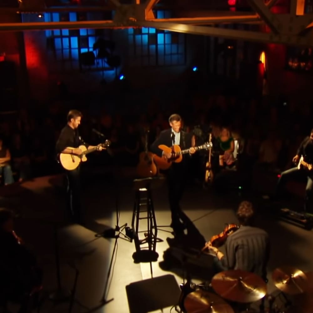 Live music performance with acoustic guitars, tambourine, drum set in an indoor venue with dim lighting and stage focus on performers.
