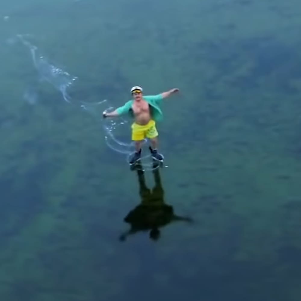 A man wearing a green sleeveless shirt, yellow swim trunks, and white cap with sunglasses stands near water at an outdoor location during daytime.