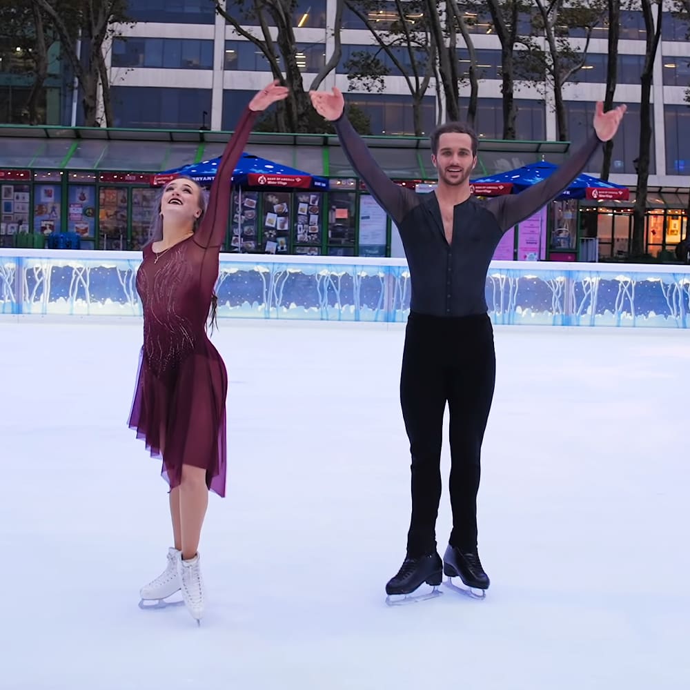 A young adult female figure skater in a maroon dress with embellishments, skating alongside an adult male at an outdoor ice rink near a kiosk displaying various items for sale. The setting is late afternoon or early evening with trees and buildings in the background.