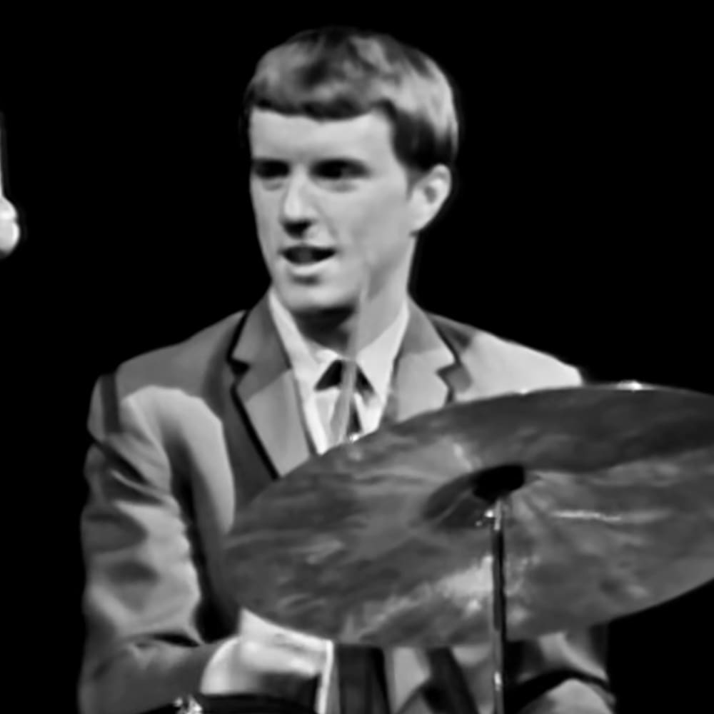 A young adult male playing drums in an indoor setting with a black background, wearing a light-colored tailored suit.
