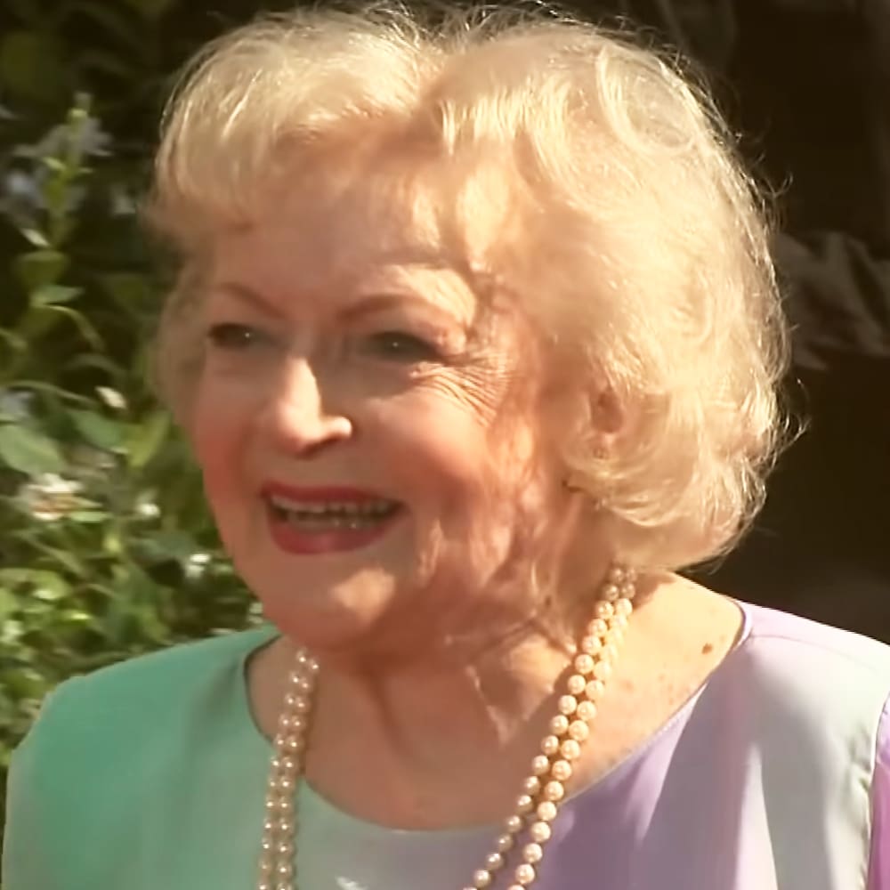 An older adult female wearing a fitted light purple and teal blouse in a garden setting with natural daylight, likely spring or summer.