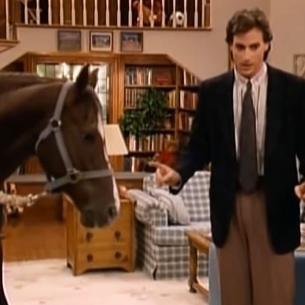 An adult male in a dark suit jacket with a young female seated behind him in front of a horse indoors.