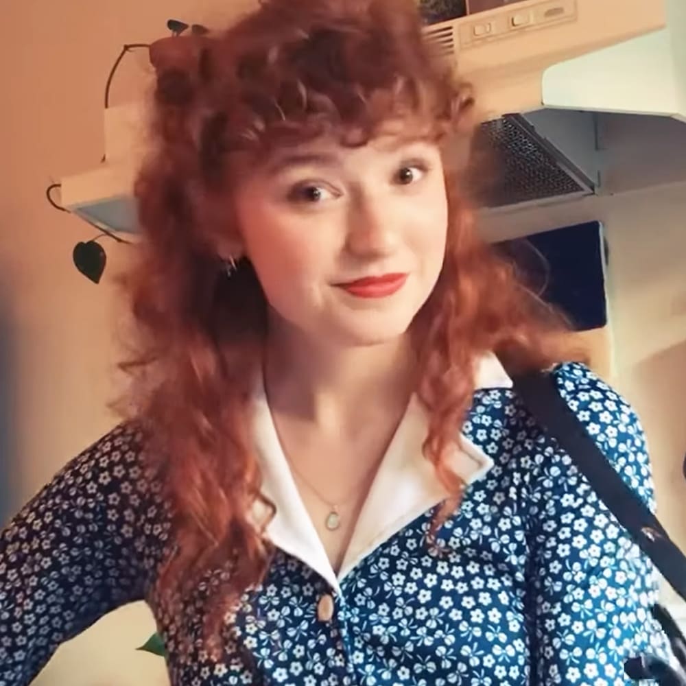 Young adult female wearing a relaxed, slightly oversized button-up shirt with a floral print in an indoor setting. Glasses hanging on the wall and greenery are visible in the background.