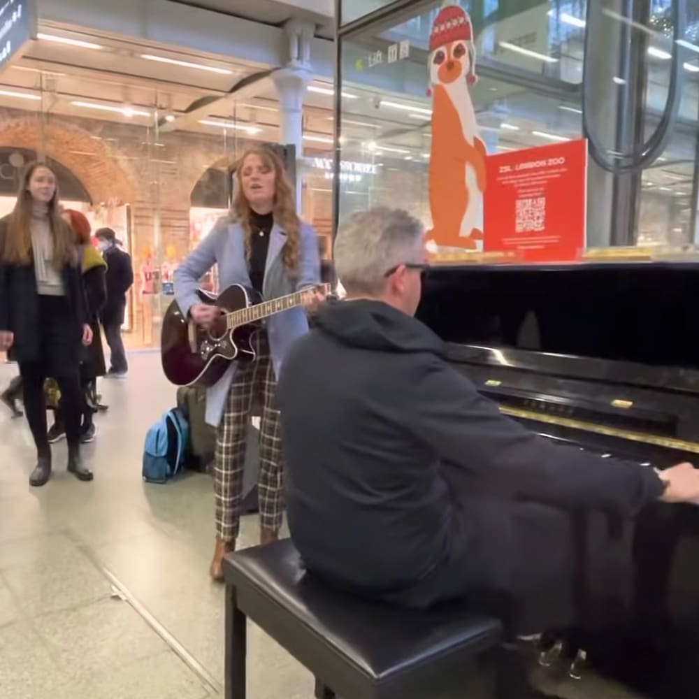 A woman playing guitar in an indoor public space with a piano bench, glass walls, brick arches, and other people walking by. The setting suggests daytime with artificial lighting.