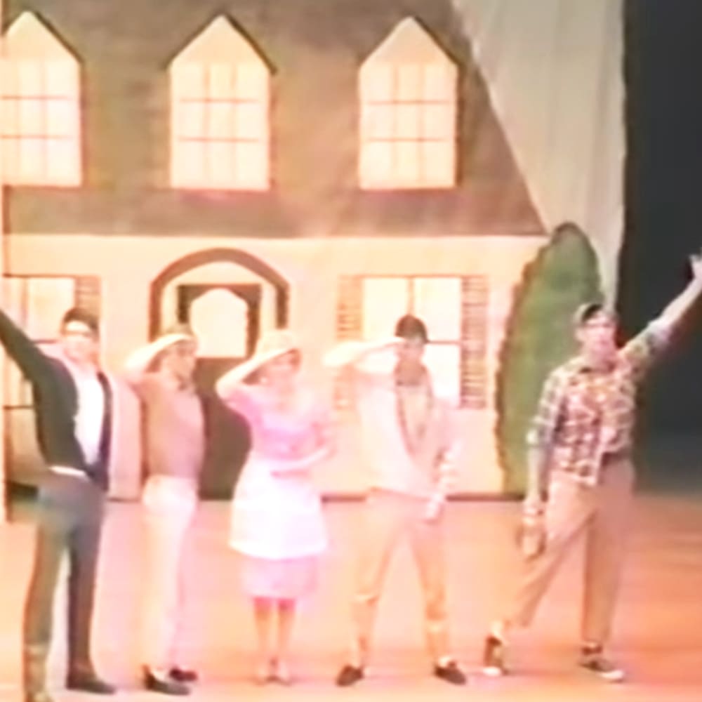 A theatrical performance on an indoor stage with characters dressed in period attire representing a home or village scene from a play.
