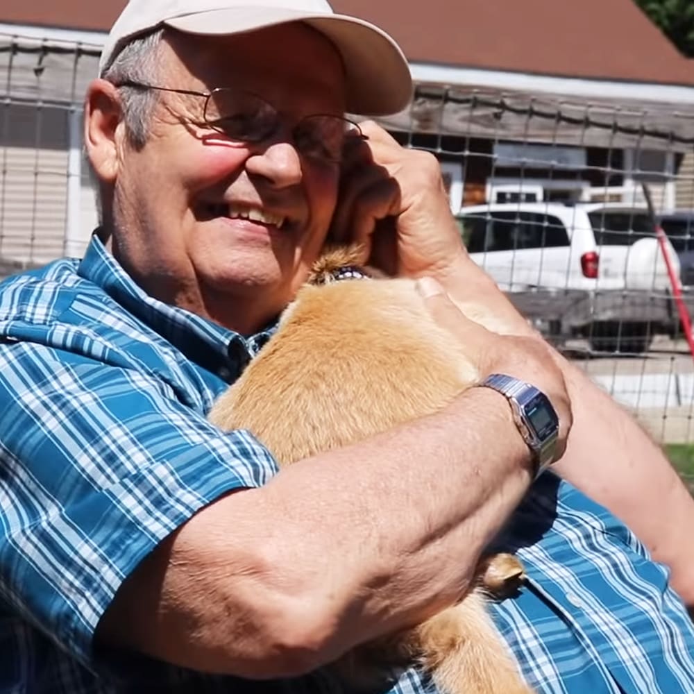 An older adult male holding a small dog in front of him with a white pickup truck and chain-link fence visible behind them.