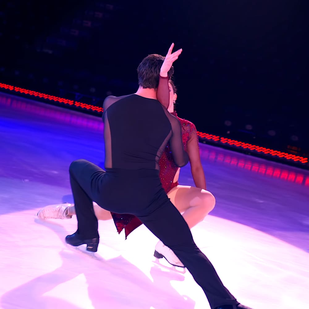 A figure skating pair performs on an indoor ice rink under stage lights, with the male skater in a black sleeveless leotard and pants, and the female skater in a red sparkly dress.