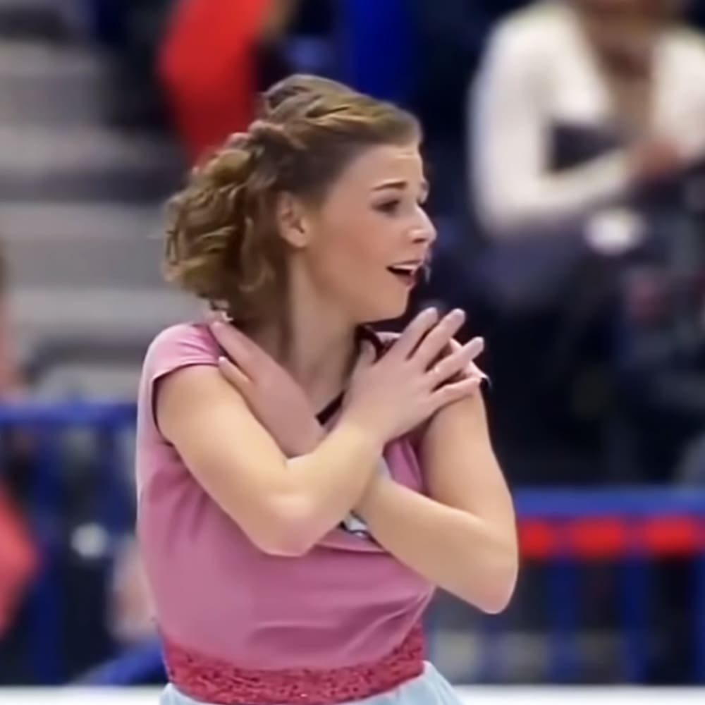 A young adult female wearing a pink, short-sleeved lace-detailed top at an indoor sports arena with blurred spectators in the background.
