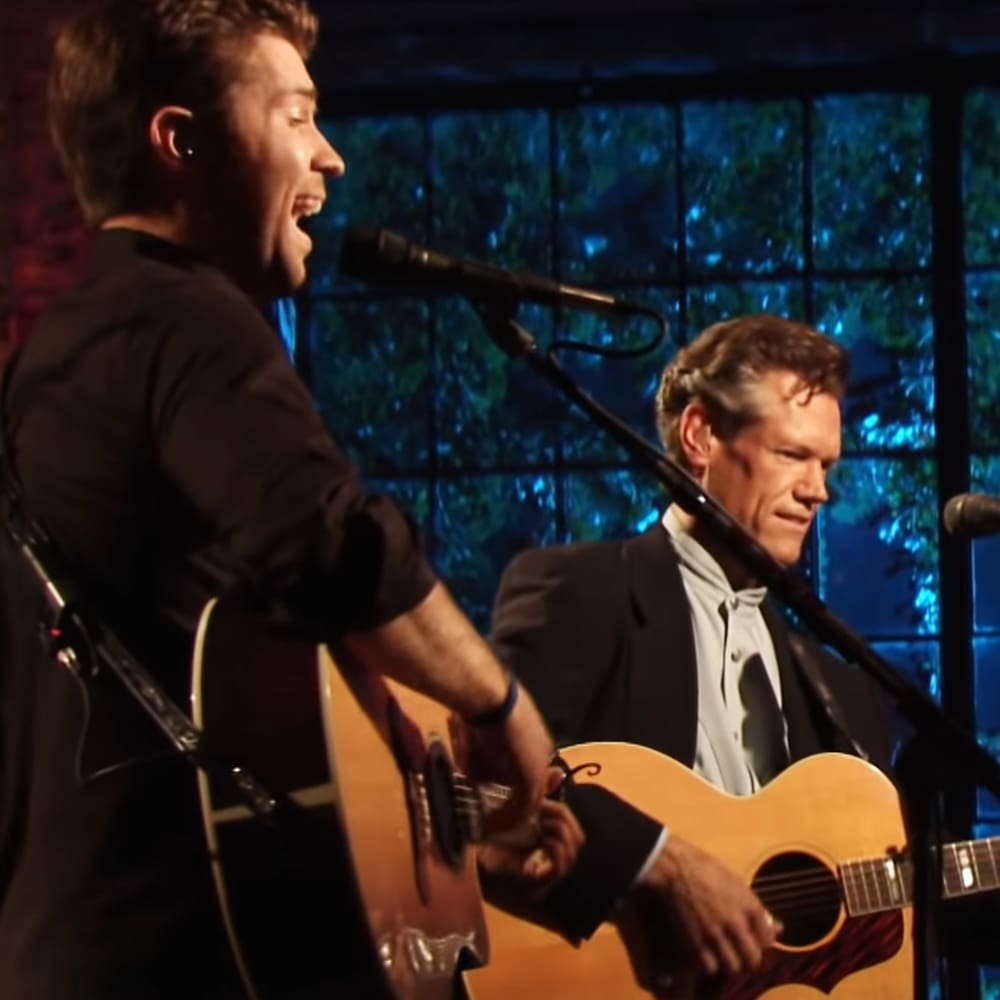 A young adult male singer with a black long-sleeved shirt, accompanied by an older male performer in a dark suit jacket and white button-up shirt, both playing acoustic guitars on stage against a stained glass backdrop under artificial lighting.