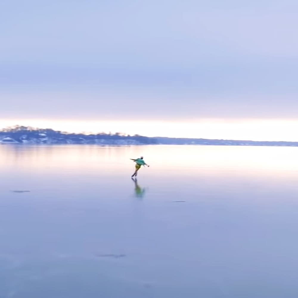 Young adult male skating on ice at a frozen lake during early morning or late afternoon, soft blue and pink hues in the sky reflecting off pale blue icy surface.