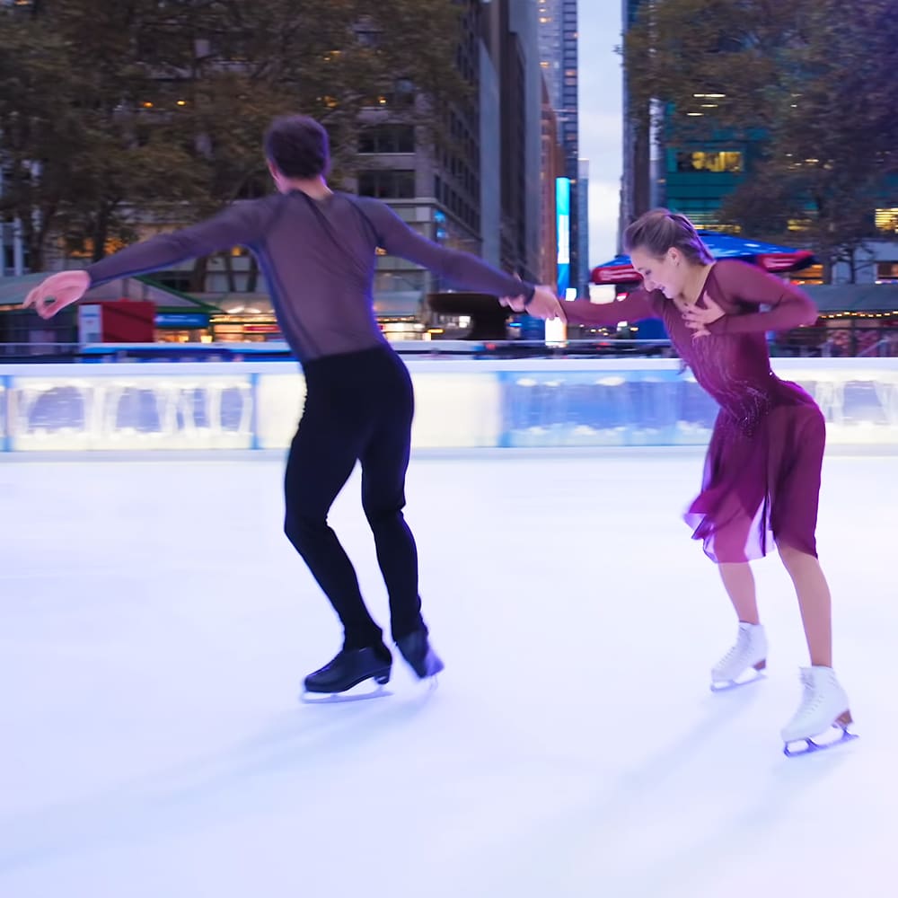 A young adult male and female ice skating in an urban outdoor setting with muted autumn colors.