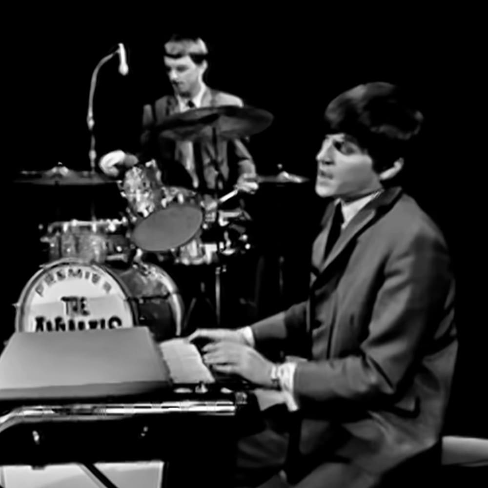 A young adult male playing an electric keyboard with a drum set in the background at what appears to be a music venue.