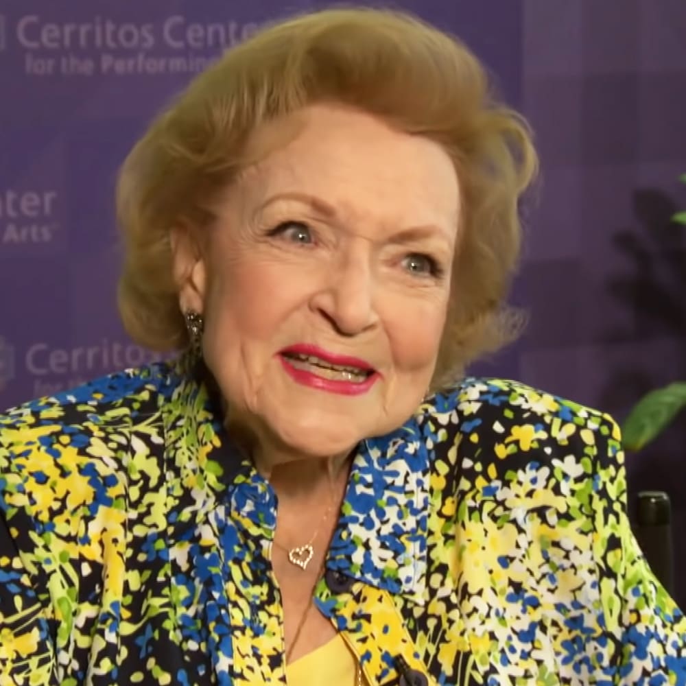 An older adult female wearing a vibrant floral jacket at a performance center with 'Cerritos Center for the Performing Arts' backdrop.