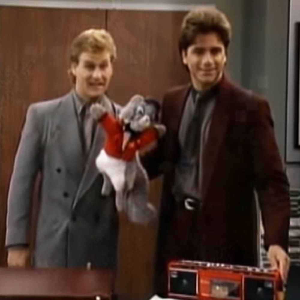 An office environment with a young adult male wearing a dark gray double-breasted suit, another in maroon, both sitting at a desk. A stuffed cat toy resembling Tom from 'Tom & Jerry' is also present.