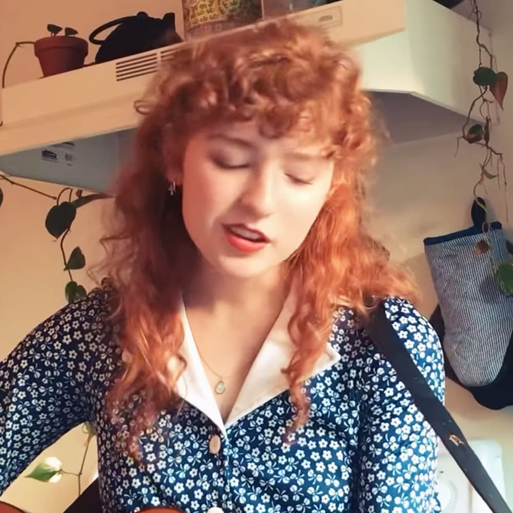 Young adult female wearing a blue button-down blouse with small white flower print, accessories include necklace and earrings. Indoors, possibly in a home or apartment setting with shelves of items and plants.