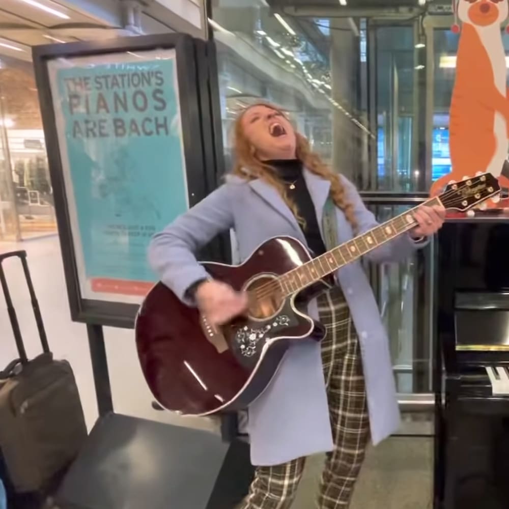 A young adult female sings while playing a guitar indoors at 'The Station's Pianos Are Bach'. She wears a light blue blazer, black top, and plaid pants.