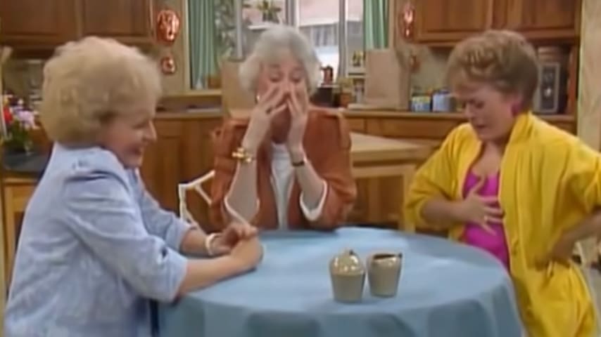 Three older adult females in a home setting, wearing casual clothing with light blue shirts and brown jackets. The background includes wooden cabinets, shelves, and kitchen appliances.