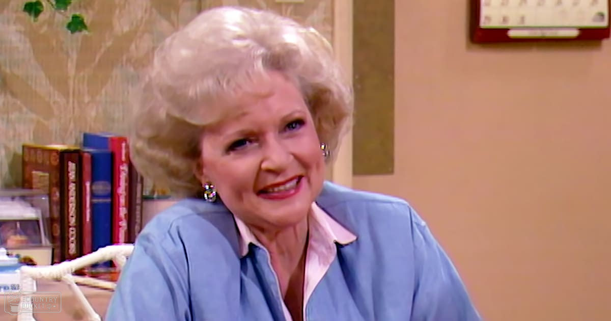 An older adult female in a relaxed, light-blue button-down shirt with dangling earrings, surrounded by books on a bookshelf and home decor elements like a calendar or picture frame within an indoor living room setting.