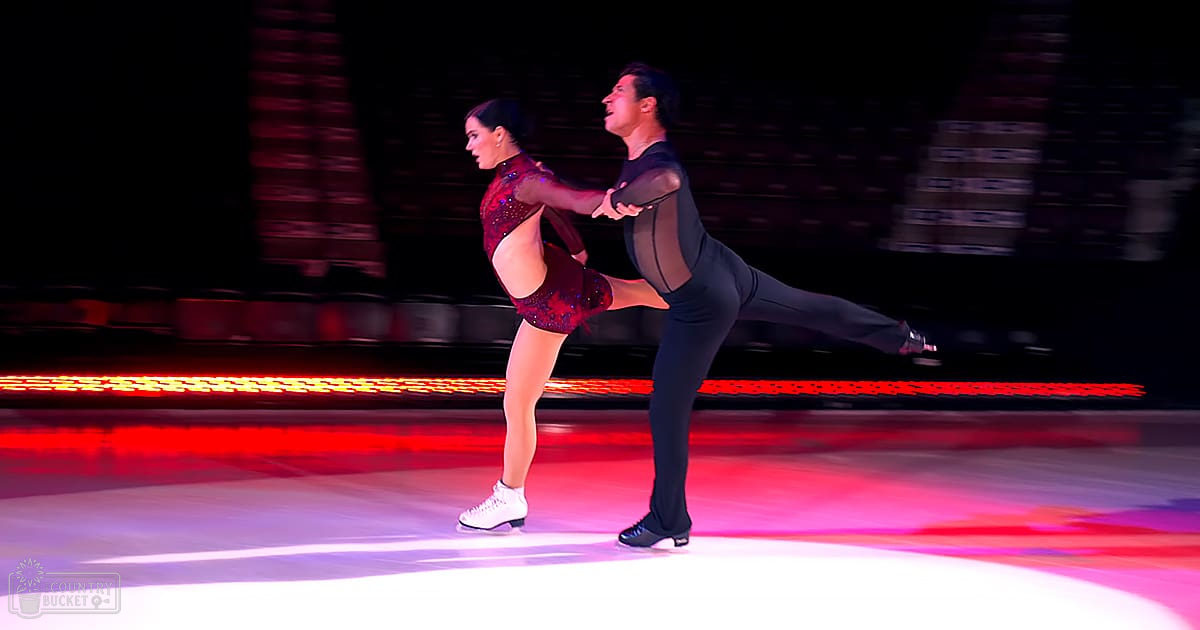 Young adult female figure skating pair performing on a professional indoor ice rink with red lighting, surrounded by blurred structures and equipment.