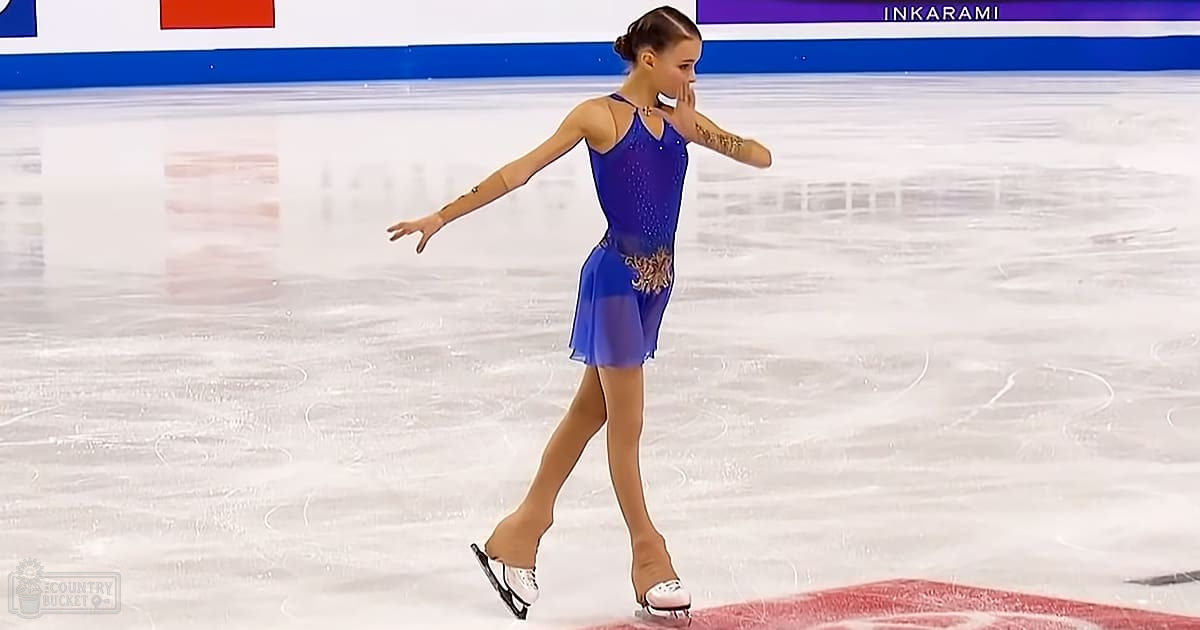 A young adult female figure skater in a blue and gold embellished dress with white figure skates on an indoor ice rink, surrounded by a blue and white barrier.