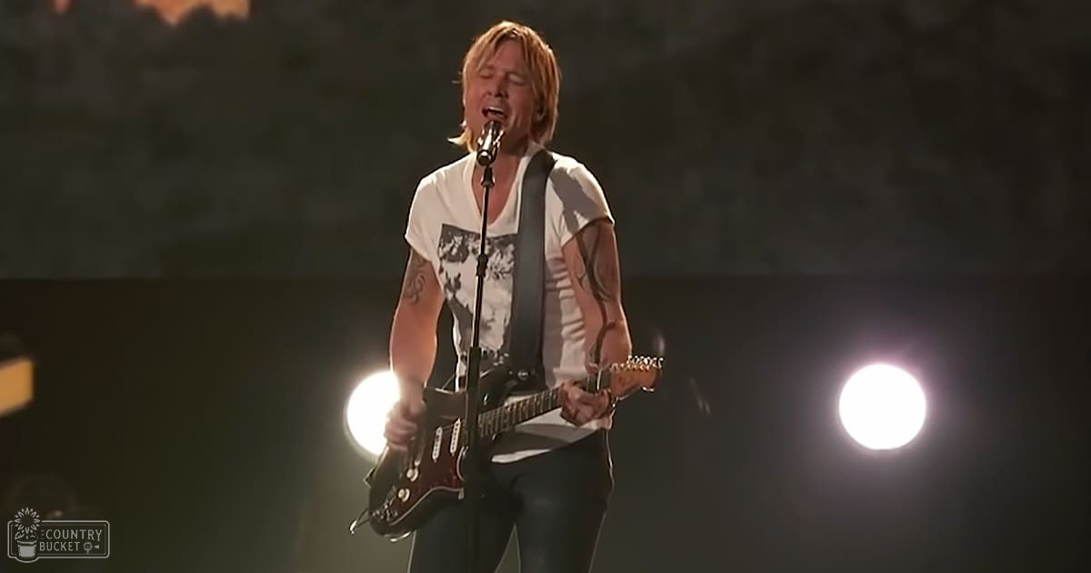 An adult male singer performing on stage, wearing a white t-shirt with graphic design and black pants, holding a guitar over his shoulder in an indoor concert venue at night under bright stage lights.