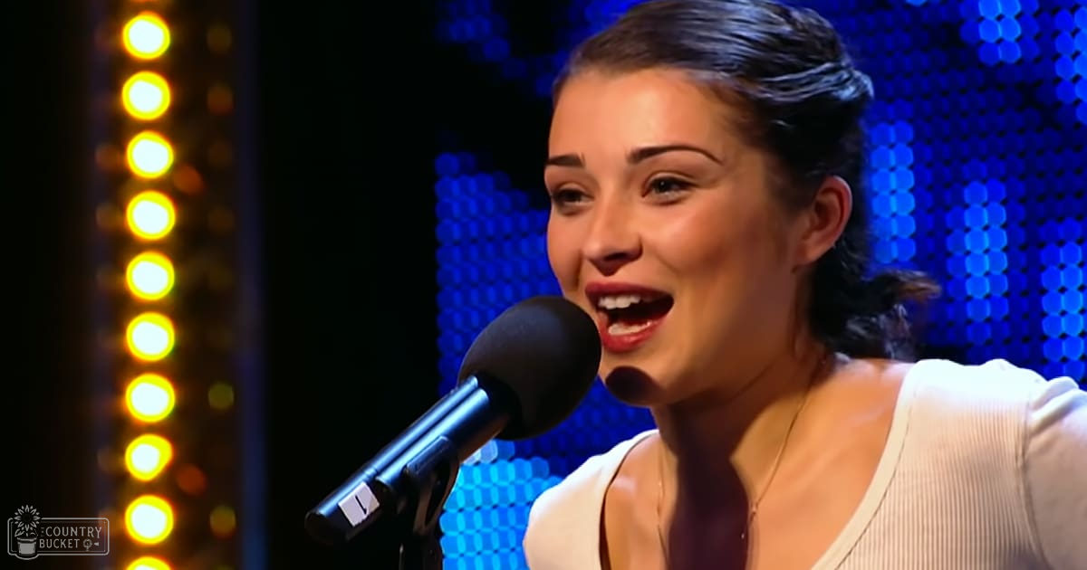 A young adult female in a white, form-fitting ribbed top with short sleeves stands on stage under bright yellow and blue circular lights. She wears a necklace.