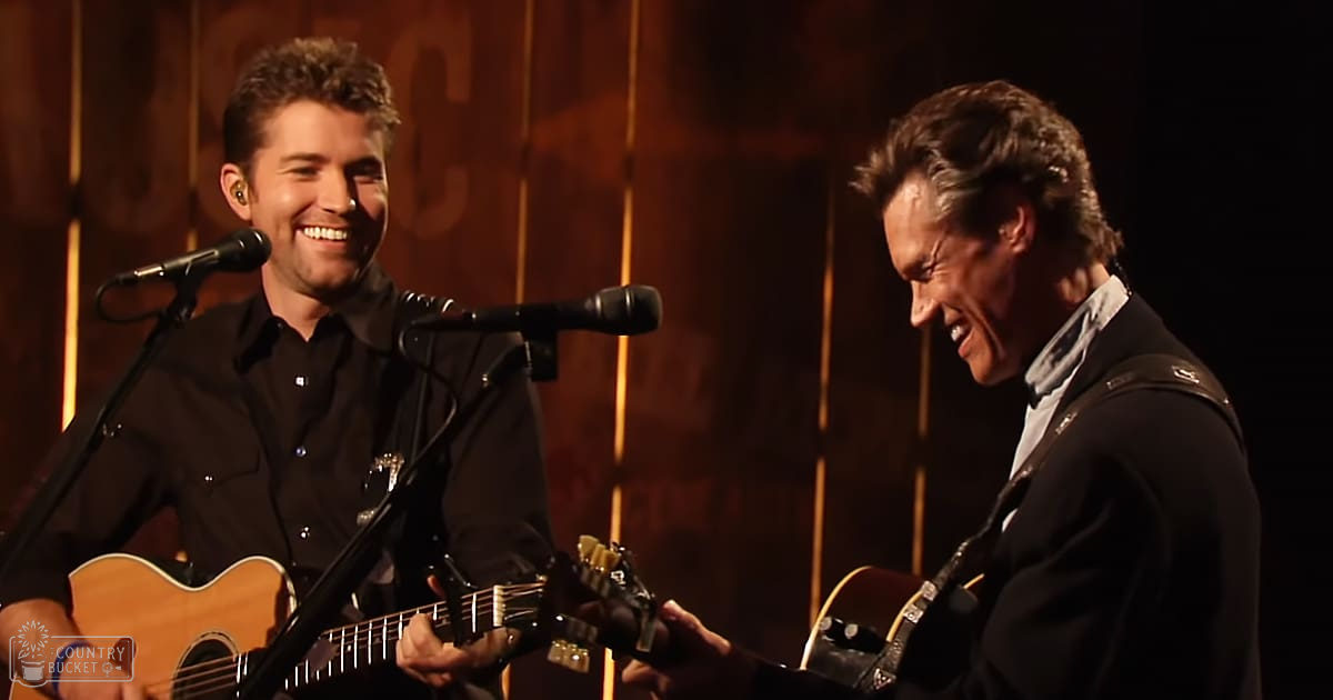 Musical performance with a young adult male singer in a black button-up shirt and an older male performer in a dark suit jacket, set against a wooden backdrop with vertical panels.