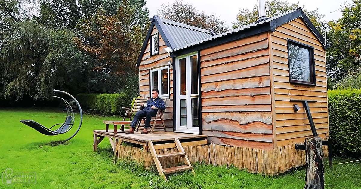 An adult male sits outside a small wooden cabin in an autumn garden, wearing a dark blue zip-up jacket and barefoot with a watch on his left wrist.