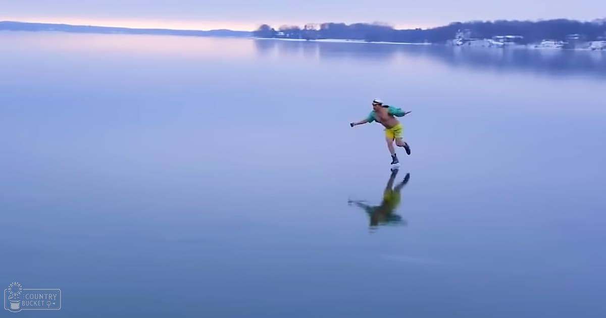 A man wearing a green t-shirt and yellow shorts stands by a serene lake under an overcast sky with reflections of trees in the water.
