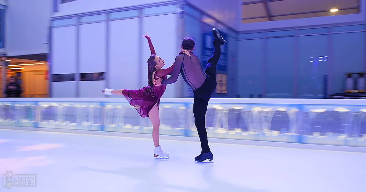 A figure skating pair performing on an indoor ice rink, surrounded by bright artificial lighting and reflective glass panels.