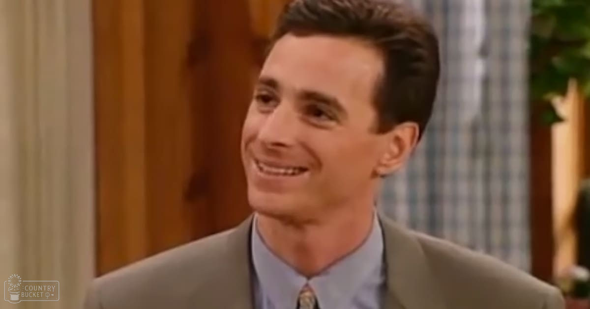 A young adult male wearing a light brown suit jacket, light blue dress shirt with a patterned tie, in an indoor setting with natural daylight and curtains.