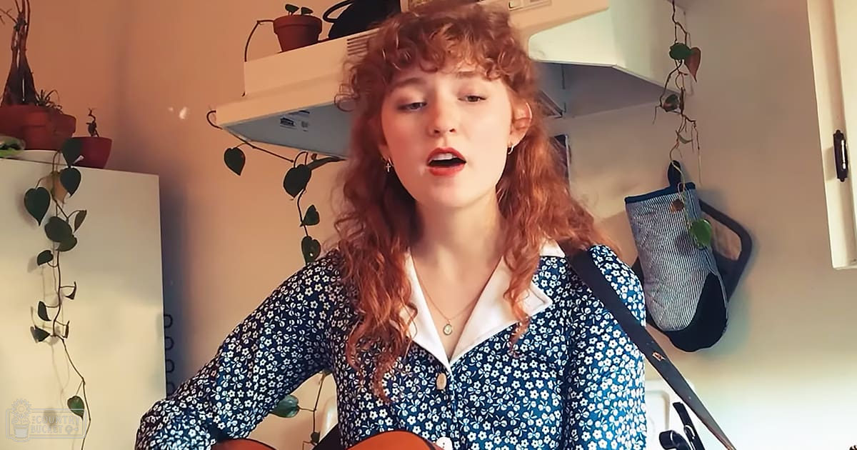 A young adult female sings in a kitchen with white shelves holding pots, plants, and utensils; greenery hangs on the wall.