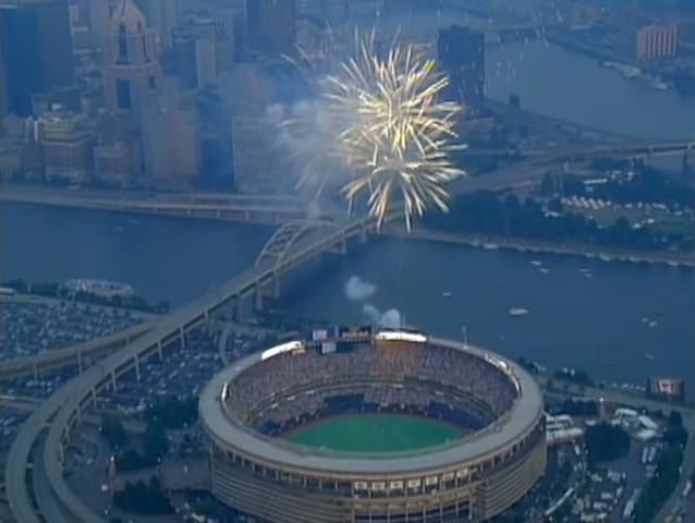 A bustling baseball stadium filled with spectators under a dramatic evening sky with a river, bridge, and cityscape in the background; fireworks explode above during what appears to be a special occasion or holiday celebration.