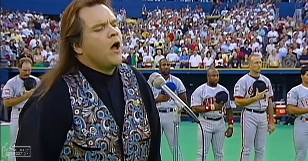 Adult male singing at a baseball stadium during daylight, with young adult and middle-aged baseball players in white uniforms in the background.