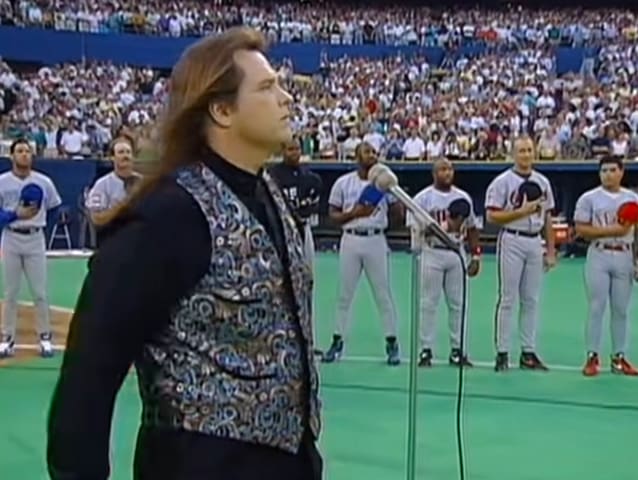 An adult male singer with long hair, wearing a black patterned vest over a white shirt, holding a microphone on a baseball field during a game.