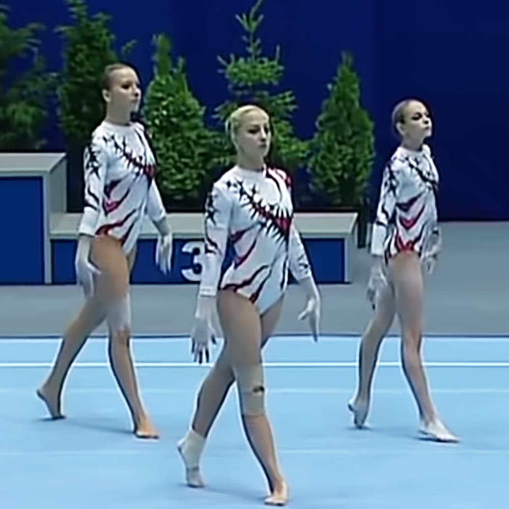 A synchronized gymnastics performance by young adult female athletes in an indoor arena with a blue wall and competition number visible.