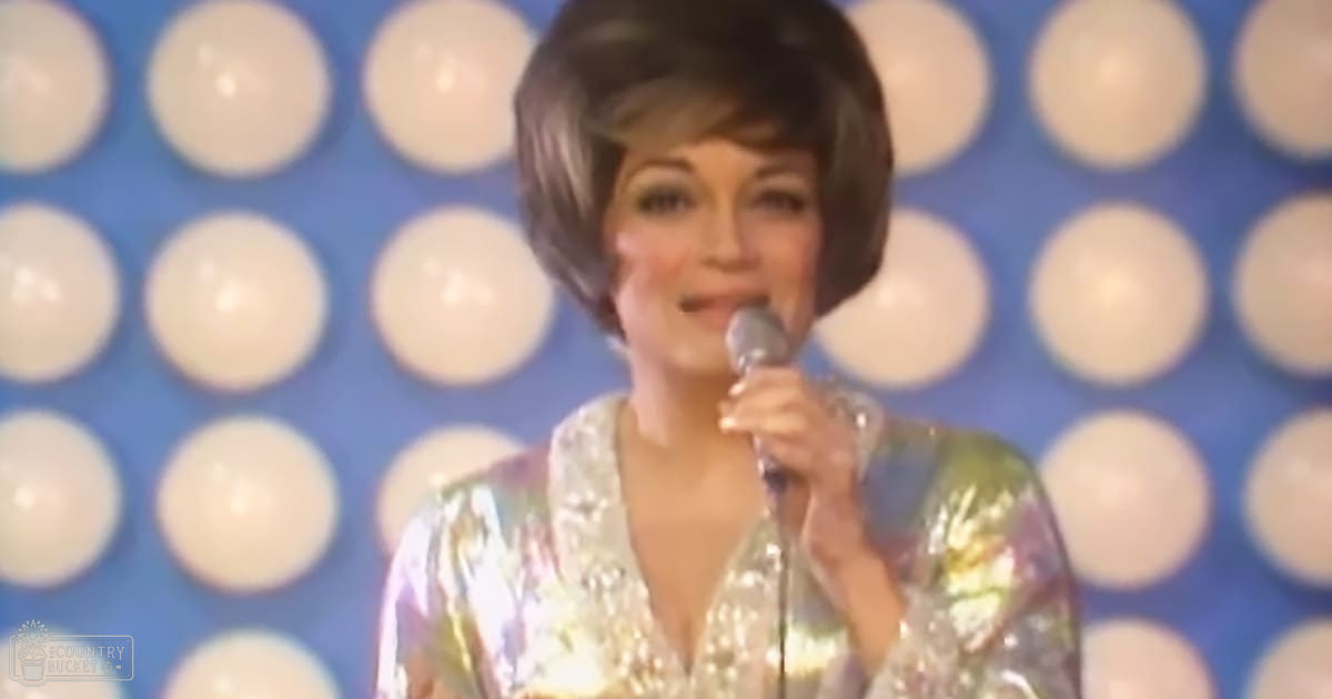 A middle-aged female performer in a shimmering sequined dress, holding a microphone against an indoor stage backdrop with blue walls and circular light elements.