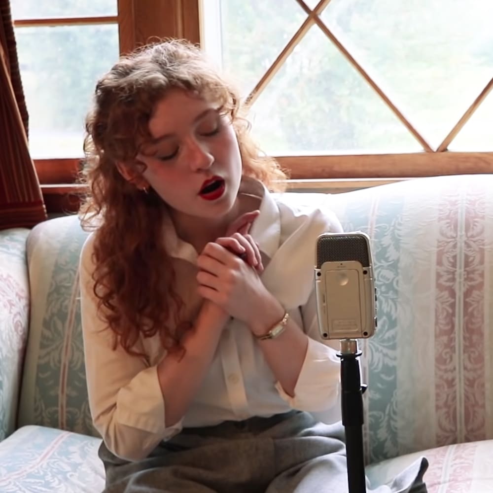 Young adult female in a casual vintage-inspired white button-up shirt with long sleeves, wearing a bracelet and red lipstick. The setting is an indoor living room or studio with natural light from large windows showing greenery outside.